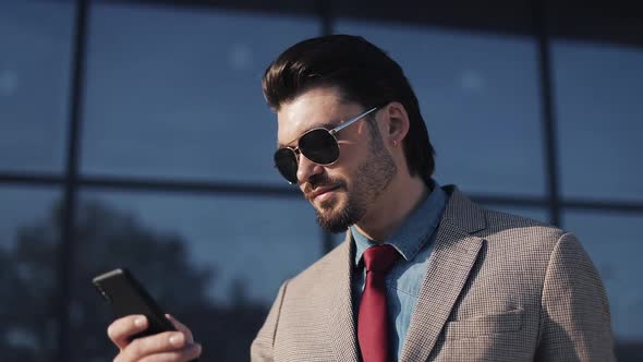Young Bussinessman in Classical Suit Wearing Sunglasses Using His Smartphone and Smiling on the alt