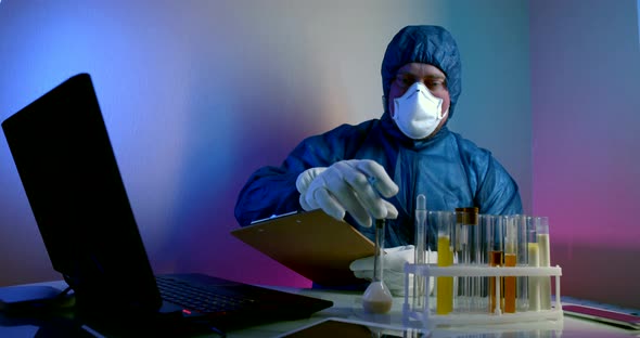 Man in Protective Clothing with a Hood, Gloves and a Respirator Is Sitting at a Table with Test alt
