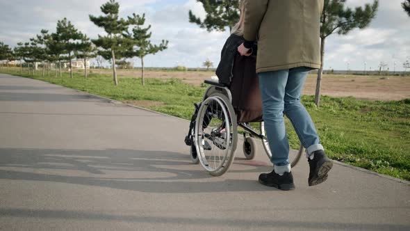 Male Is Rolling Invalid Chair with Young Sick Woman Outdoors Back and Side View alt