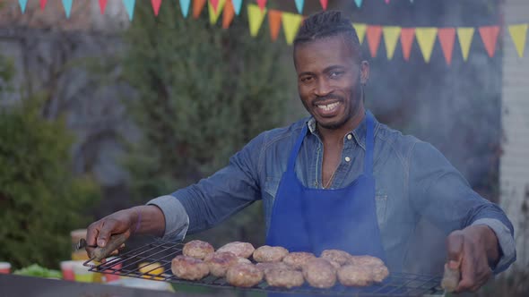 Portrait of Happy Smiling African American Man Posing with Burger Patties on Bbq Grid Outdoors alt