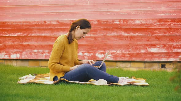 A girl with a laptop on her lap is sitting on a blanket on the lawn and typing on the keyboard alt
