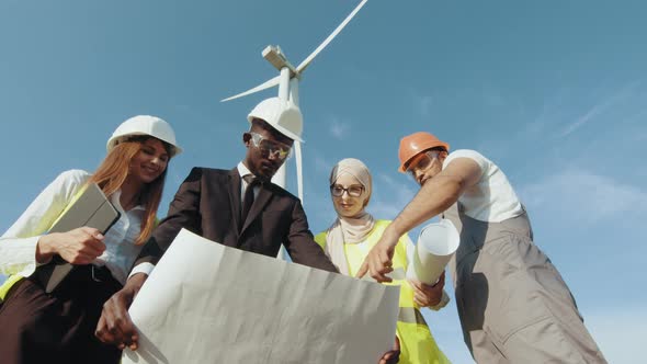 Four Multi Ethnic Partners in Safety Helmets Studying Wind Turbine Blueprints alt