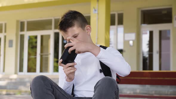 A Caucasian Teenage Boy Plays a Game on a Smartphone As He Sits in Front of School  Closeup