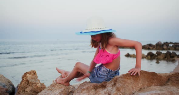 Happy Funny Teen Girl in Beach Clothes and Hat is Sitting on Stone on Seashore