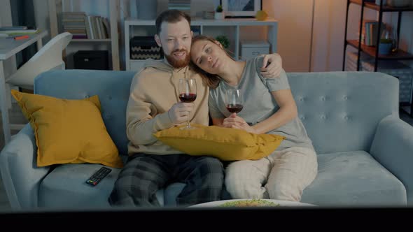 Husband and Wife Drinking Red Wine From Glasses Talking and Watching Film on TV in Apartment at