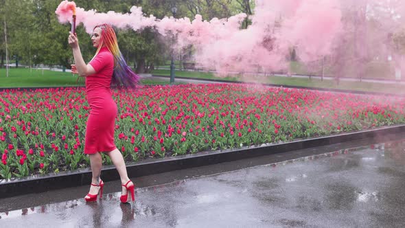A Girl with Makeup with Rainbow Braids in Red Dress Posing in Red Smoke Against the Background of a alt