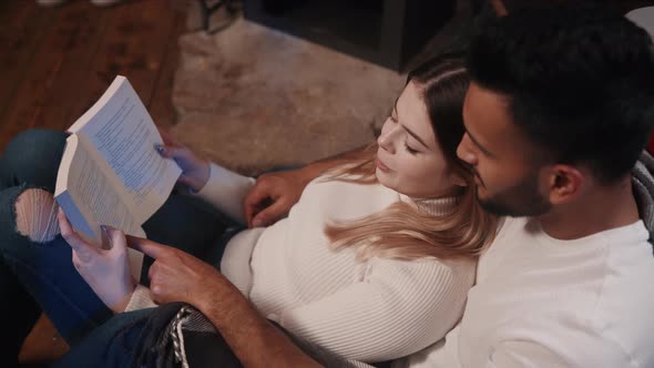 Happy Young Multiracial Couple Hugging and Reading Book Together Near the Fireplace alt