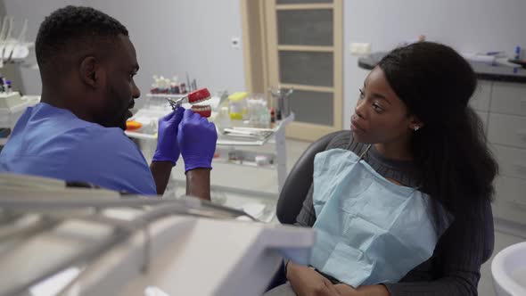 Young Handsome Smiling African American Male Dentist Showing Teeth Model to His Female African alt