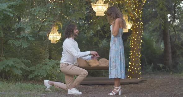 Wide Shot of Loving Romantic Man Standing on One Knee and Proposing To Beautiful Young Woman alt