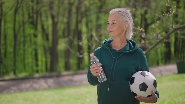 Live Camera Zoom in to Smiling Satisfied Senior Caucasian Sportswoman Standing with Soccer Ball and alt