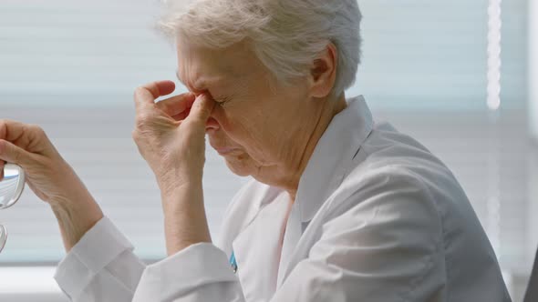 Tired Senior female doctor in coat massages temples suffering from headache alt
