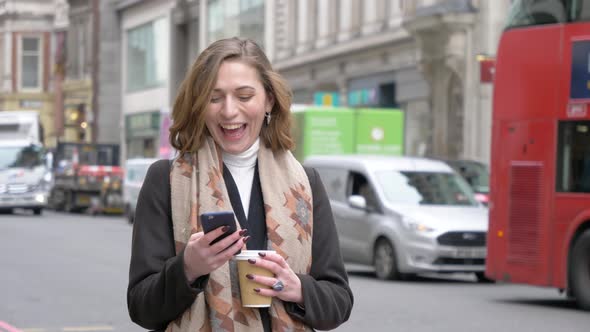 Laughing blond woman using smartphone and holding coffee cup in London alt