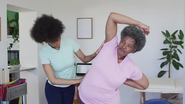 Happy african american female physiotherapist helping senior female patient exercise at home alt