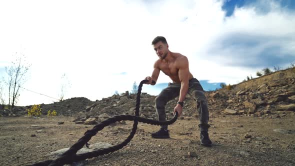Muscular athlete doing waves with battle ropes in nature. Professional sportsman exercises with cabl alt