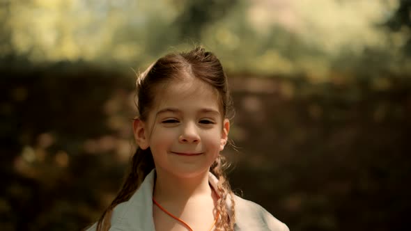 Cute Little Girl Portrait Smiles On Nature Looking At Camera On Botanical Garden. Funny Cheerful Kid alt