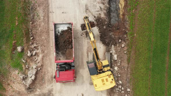 Top View Yellow Excavator Picks Up Land From the Field and Loads It Onto a Red Truck alt
