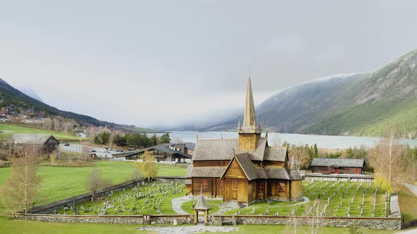 Stave Church And Graveyard Overlooking Otta River And Snowy Mountain In Lom, Norway. - aerial alt