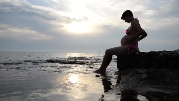 Pregnant Woman In A Swimsuit Sitting In Profile Cradling Her Swollen Belly alt
