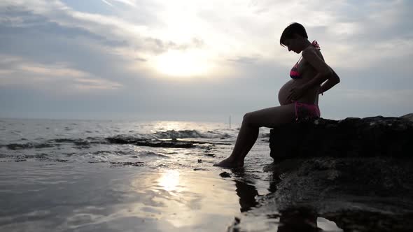 Pregnant Woman In A Swimsuit Sitting In Profile Cradling Her Swollen Belly