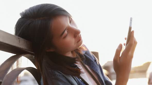 Sad girl using a smartphone while sitting beside a river in the sun at sunset.