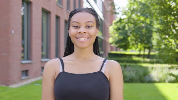 A Young Black Woman Smiles at the Camera - an Office Building and Green Park in the Background alt