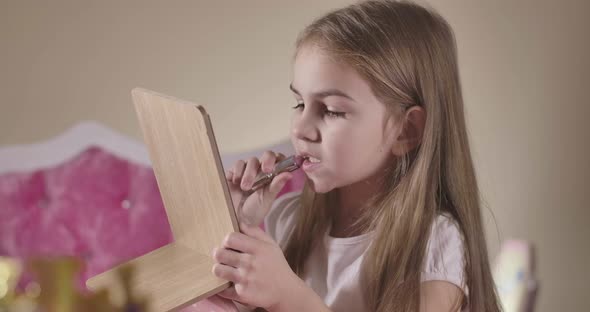 Cute Caucasian Girl Sitting in the Bedroom and Applying Red Lipstick. Pretty Child Taking Care of alt