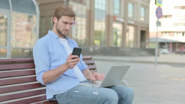 Man Using Smartphone and Laptop While Sitting Outdoor on Bench alt