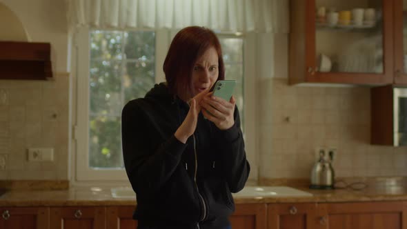 Young Adult Woman Reads Good News on Cell Phone Standing in the Kitchen