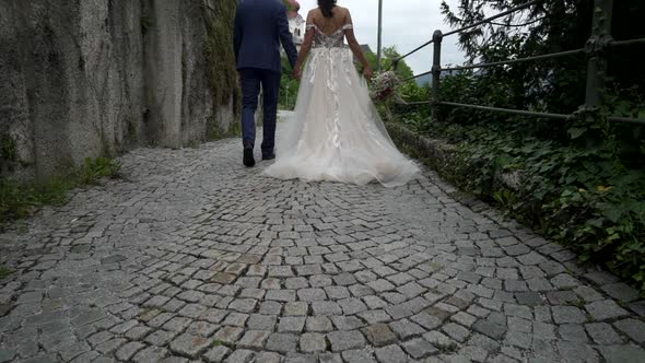 Wedding couple groom, bride walk by hand along stone wall with plants, lanterns near chapel Salzburg