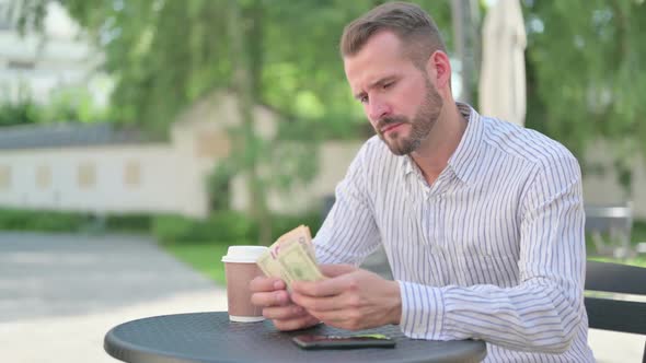 Middle Aged Man Counting Dollars While Sitting in Outdoor Cafe alt