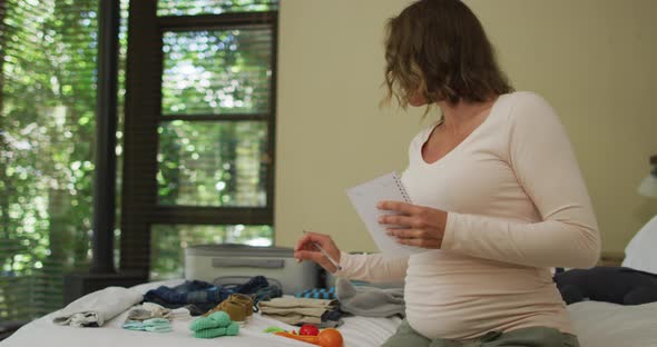 Caucasian pregnant woman holding list and packing suitcase to hospital for delivery alt