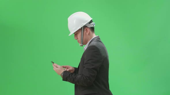 Side View Of Asian Chief Engineer In The Hard Hat Use Mobile Phone While Walking On Green Screen alt