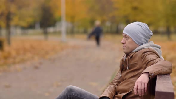 A bored young man in a gray hat is resting while sitting on a bench in an autumn park, copyspace alt
