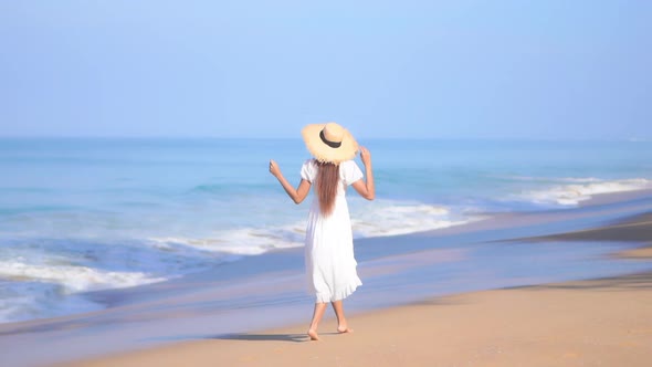 Back View Of Young Woman in White Sundress Walking Along the Sandy Beach by the Sea alt