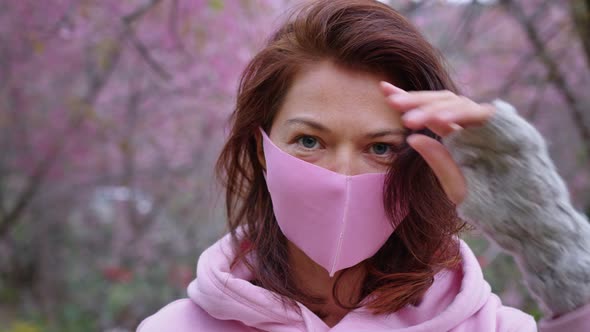 a Woman in a Pink Hoodie and a Pink Protective Mask Stands in the Sakura Garden and Looks Directly alt