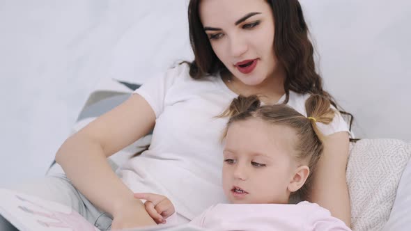 Beautiful Mother and Toddler Daughter Are in Bed Hugging and Reading a Book alt