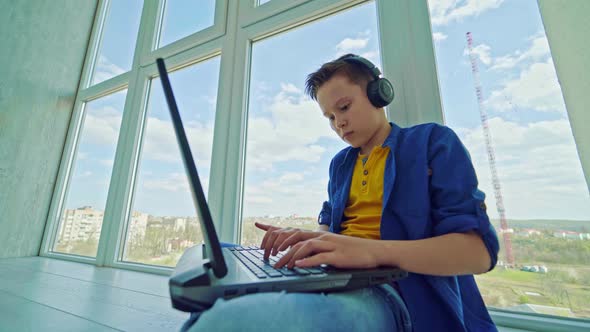Teenager in headphones with a laptop by the window alt