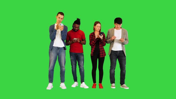 Young Friends Stand in Front of Camera Focused on Their Gadgets on a Green Screen Chroma Key alt
