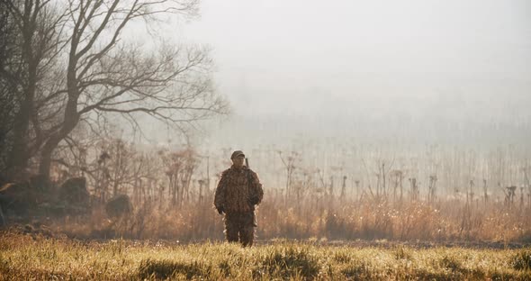 Hunter in hunting equipment with rifle on his shoulder walks through on camera in the field alt