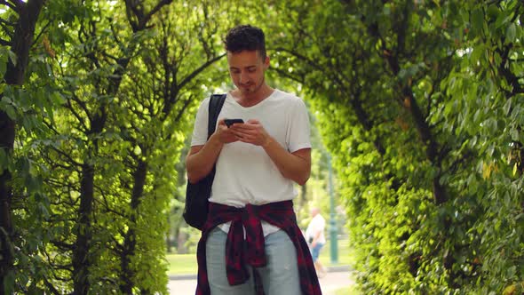 Handsome Young Man Stands in the Park Inside a Tunnel of Trees with a Phone in His Hands alt