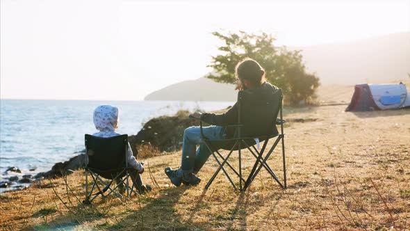 Father with Little Daughter Sits on Chairs at Camping on Lake Shore Rear View alt