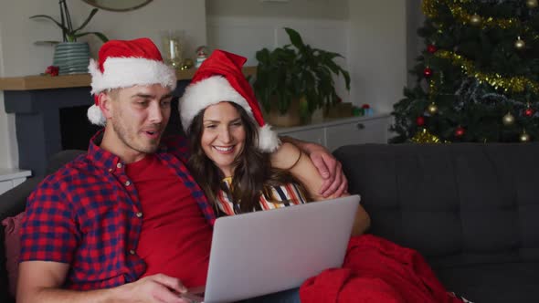 Happy caucasian couple wearing santa hats sitting on sofa and using laptop alt