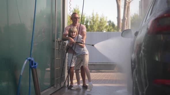 Wide Shot Portrait of Satisfied Cute Caucasian Boy Helping Man Washing Automobile at Self Service alt