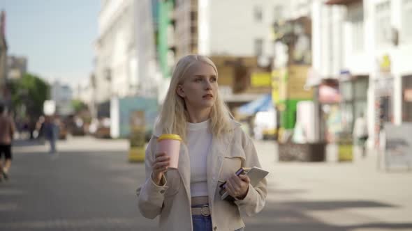 A Beautiful Young Blonde Woman Walks Along the Central Street with Books and Aromatic Coffee alt