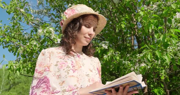 Slow Motion, Girl in Hat Sitting in Park Near a Flowering Tree Is Reading Book. Close-up, View of alt