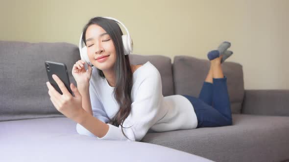 A young attractive woman lies on the couch as she listens to music from her smartphone through her B alt