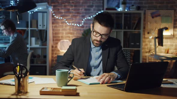 Tired Businessman Yawning Working in Office Writing in Notebook Sitting at Desk alt