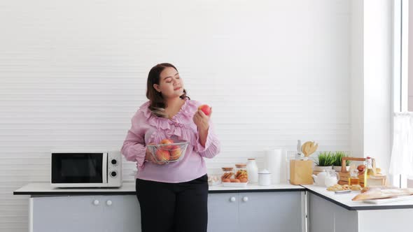 Overweight young woman standing and eating apple deliciously in the kitchen