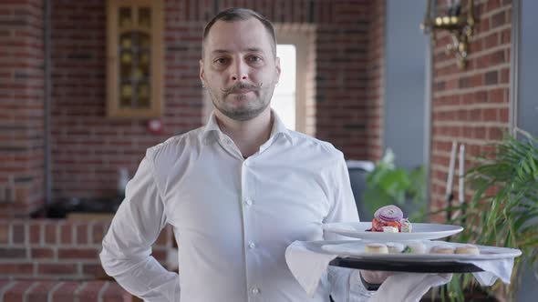 Portrait of Male Waiter Looking at Camera Walking in Slow Motion Holding Tray with Dessert and Salad alt