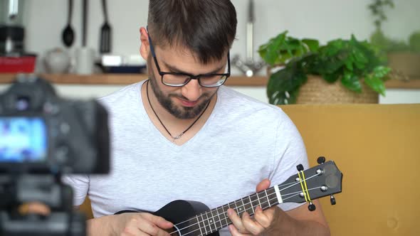 Portrait of Young Male Smiling Blogger Records Video on Camera Playing Guitar at Home alt